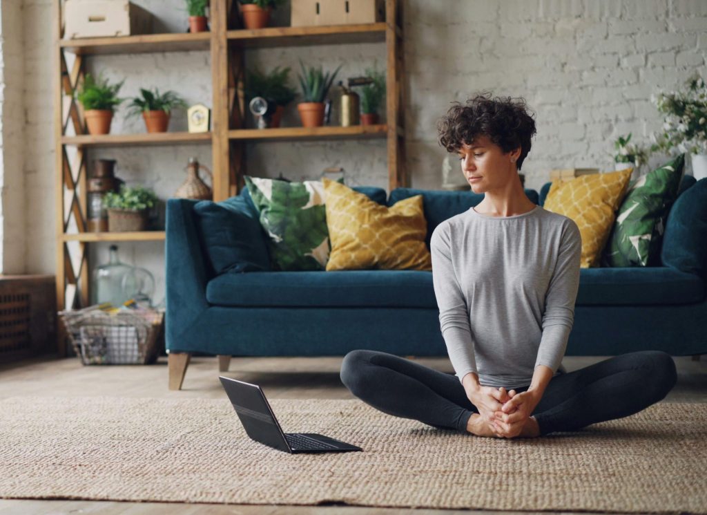 A woman meditates in a bright, cozy living room with plants and a sofa.